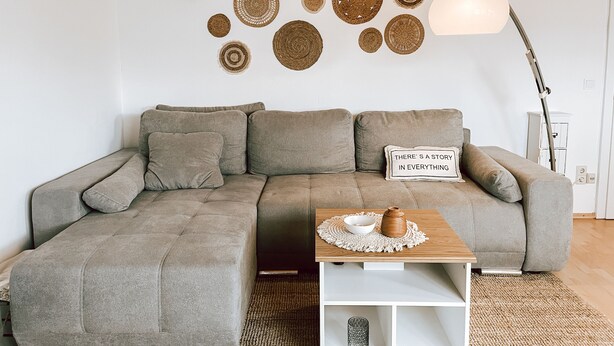 A living room with a grey sectional sofa, a small wooden side table, and a wall decorated with circular woven art pieces. - Cozy vacation apartment for 4 people (Baumholder)