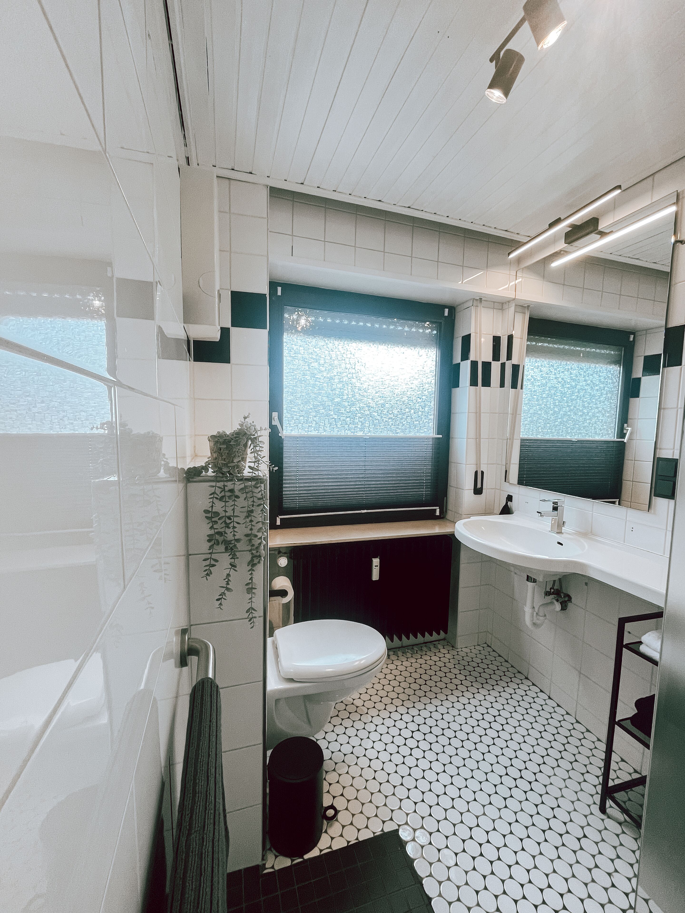Un baño con inodoro, lavamanos y bañera. Hay una ventana con persianas, un espejo y una planta. El suelo tiene un diseño de baldosas con motivos.
