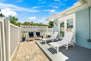 A patio area with white lounge chairs and a small table, a white fence, and a blue building with a door.
