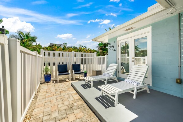 A patio area with white lounge chairs and a small table, a white fence, and a blue building with a door.