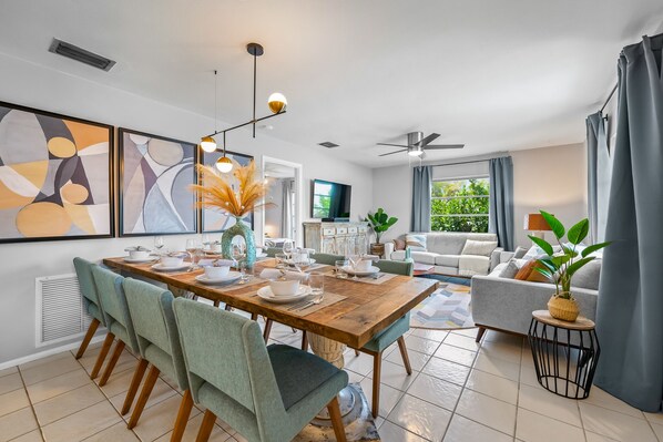 A modern living room with a dining area, featuring a wooden dining table, green upholstered chairs, a ceiling fan, and a large window with curtains.