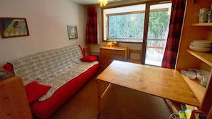 A compact room with a wooden table, a red sofa, a small desk, and a window with curtains. - Le charmant Songe en Chartreuse (Saint-Pierre-de-Chartreuse)
