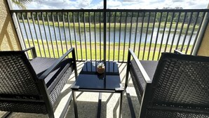 Ein Balkon mit Tisch und zwei Stühlen, mit Blick auf eine Wiese und ein Gewässer.
