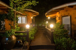 Un chemin en bois mène à un bâtiment doté d’une porte et de fenêtres, entouré de verdure et illuminé par des lumières.