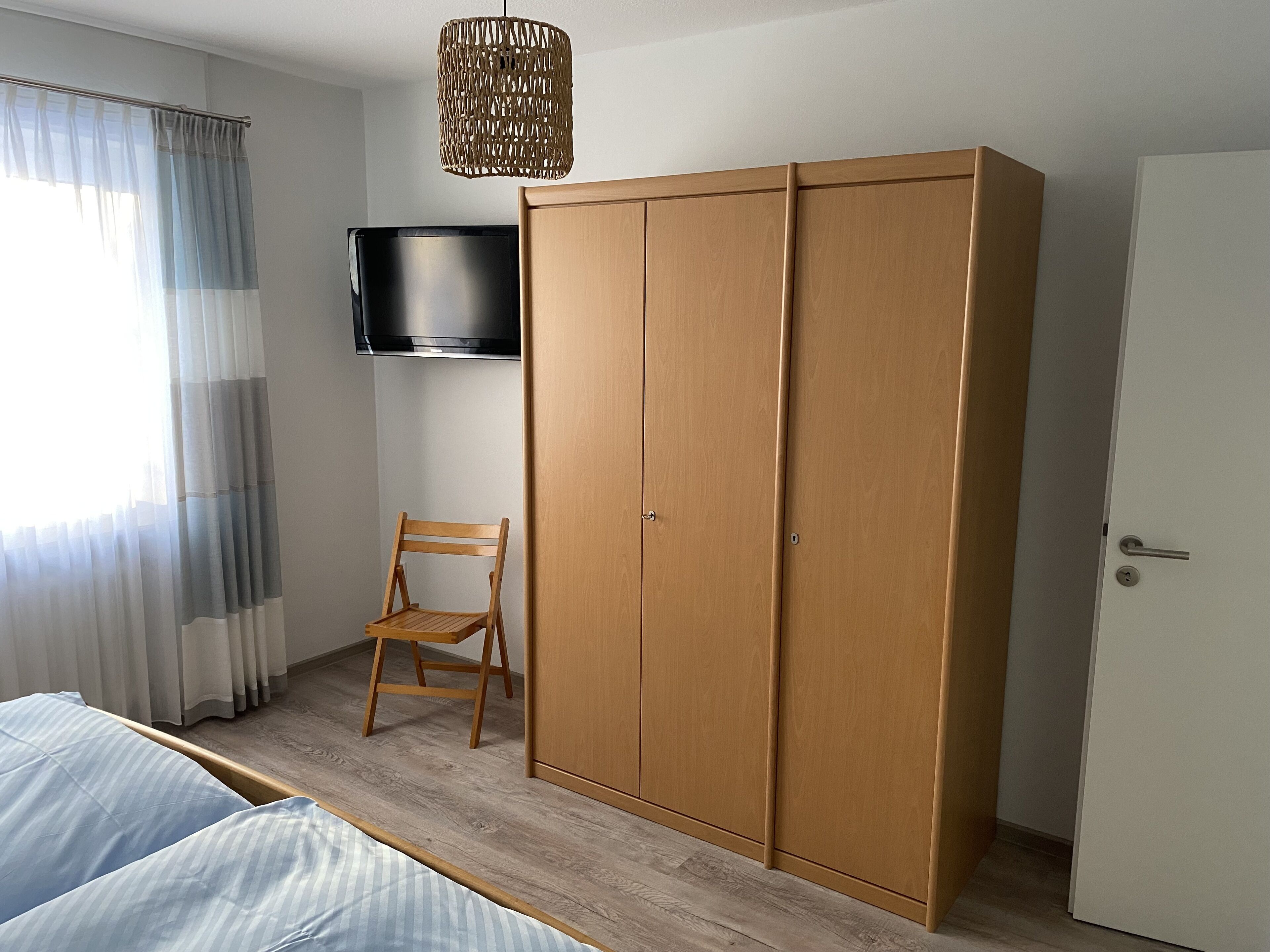 A bedroom with a bed, a wooden wardrobe, a television mounted on the wall, and a chair.