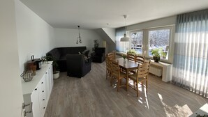 A modern living room with a black sofa, a dining table with chairs, and a large window with curtains. - Generously equipped and very spacious, family vacation apartment (4) (Lembruch)