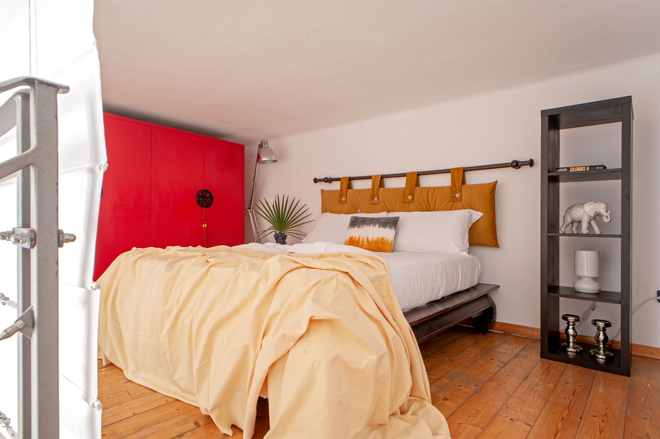 Un dormitorio con una cama, un armario rojo, una planta y una estantería con objetos decorativos.