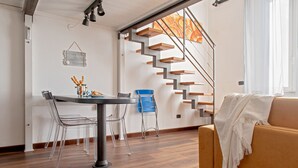 A modern living room with a wooden staircase, a dining area with clear chairs, and a sofa with a white throw.