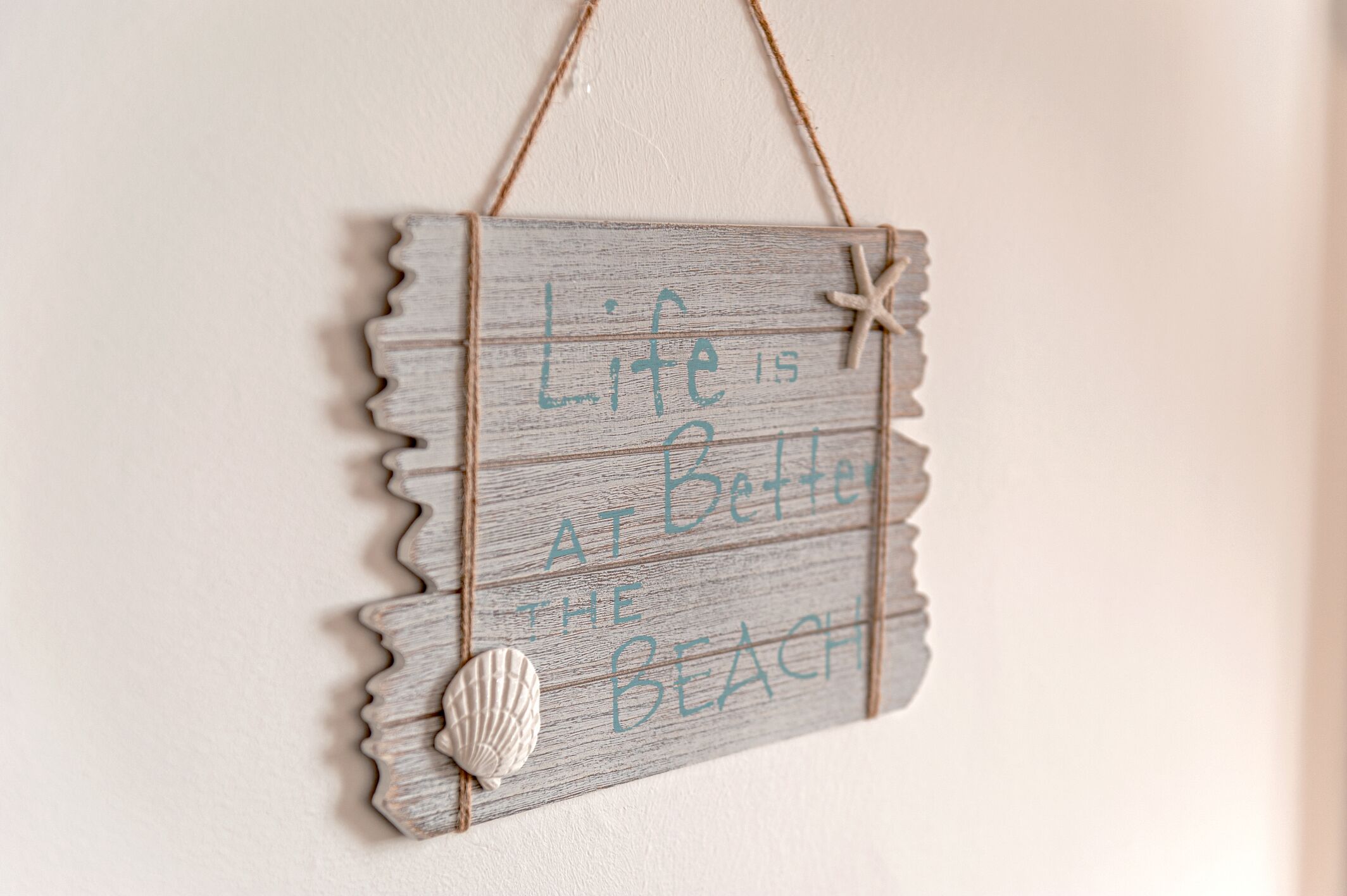 Une enseigne en bois portant l’inscription « La vie est plus belle à la plage » et ornée d’un coquillage.