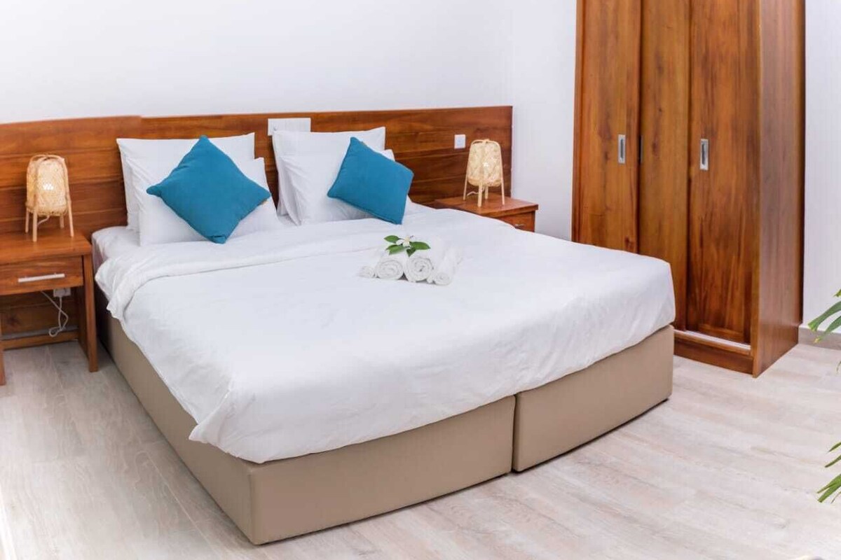 Ein ordentlich bezogenes Bett mit weißen Laken und blauen Kissen, ein Holzschrank und ein Nachttisch mit Lampe.