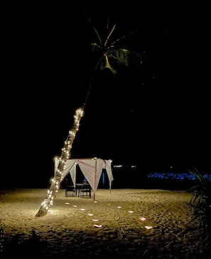 A beach tent with string lights, a table, and chairs under a palm tree at night. - Nihaali Maldives (Kudarikilu)