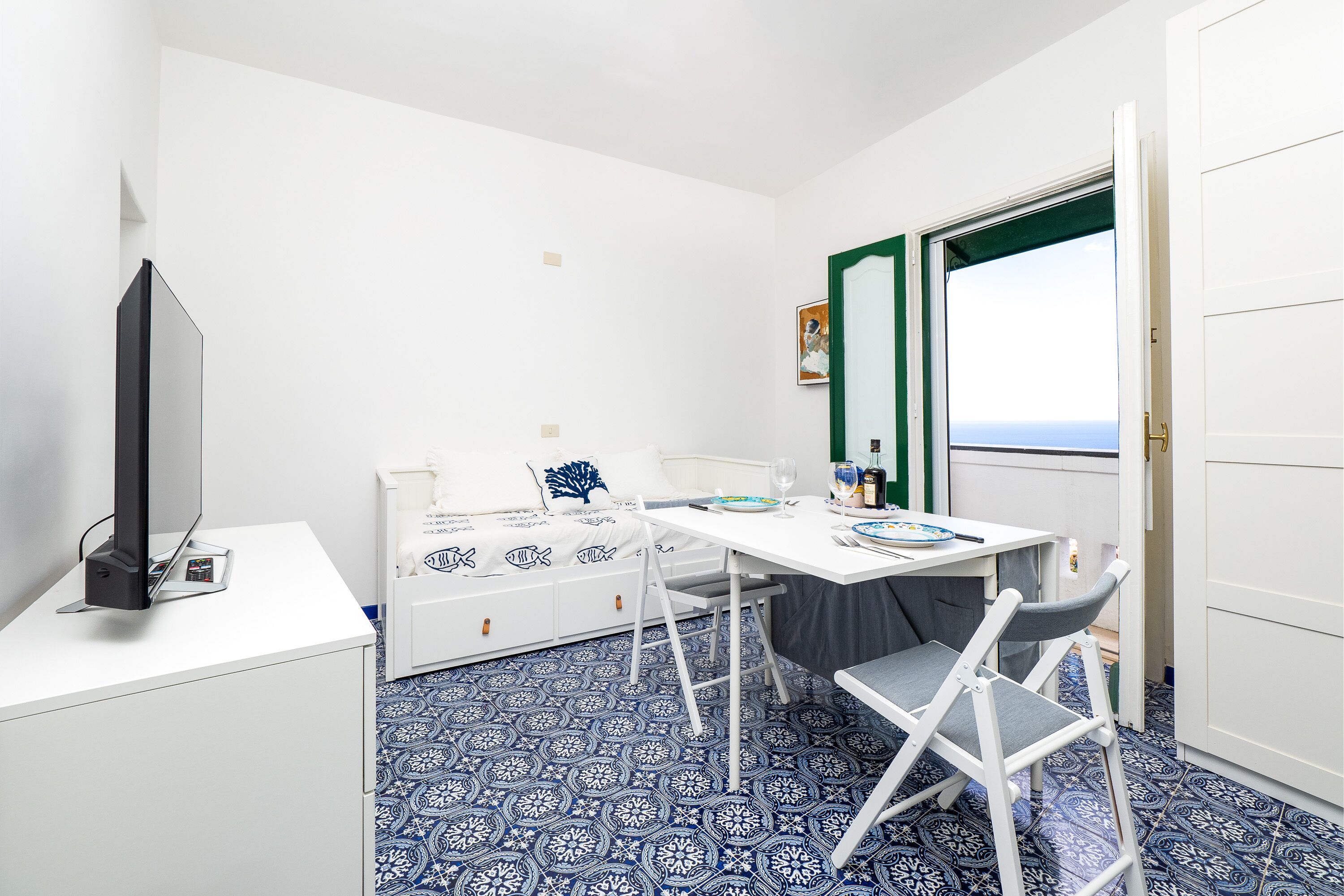 A compact room with a white bed, a small table with chairs, a television on a white cabinet, and a window with a view.