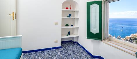 A bathroom with a blue and white patterned floor, white walls, a green-framed window, and a view of the sea.