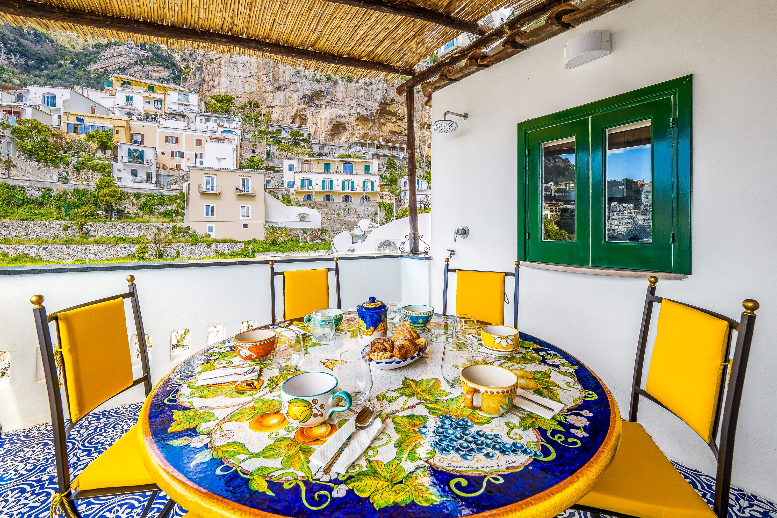 A round table set with plates, cups, and cutlery on a colorful tablecloth, overlooking a coastal village with buildings on a hillside.