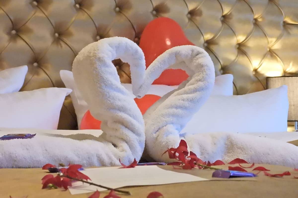 A bed with a tufted headboard, white pillows, and a heart-shaped towel arrangement.