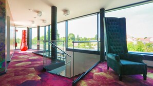 A modern hotel lobby with a staircase, a large window offering a view of greenery, and a plush armchair. - SPREE. Hotel am Hafen (Lübbenau/Spreewald)