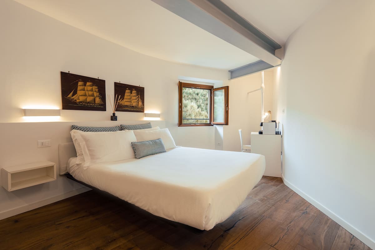 Une chambre à coucher avec un grand lit, une table de chevet, une commode, une fenêtre donnant sur un beau paysage et des œuvres d’art accrochées au mur.