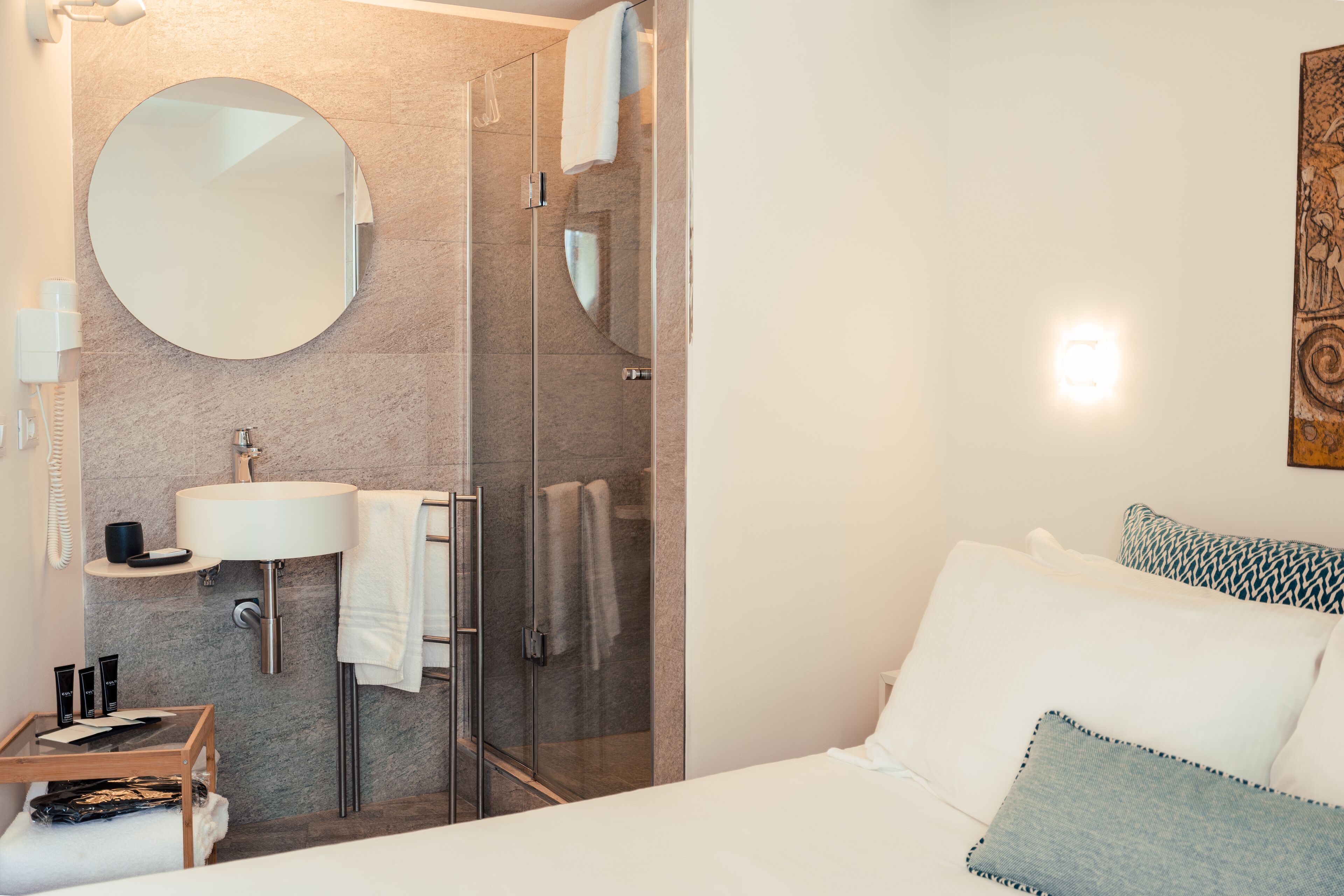A modern bathroom with a glass shower enclosure, a round mirror, a sink, and a bedside table with toiletries.