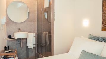 A modern bathroom with a glass shower enclosure, a round mirror, a sink, and a bedside table with toiletries.