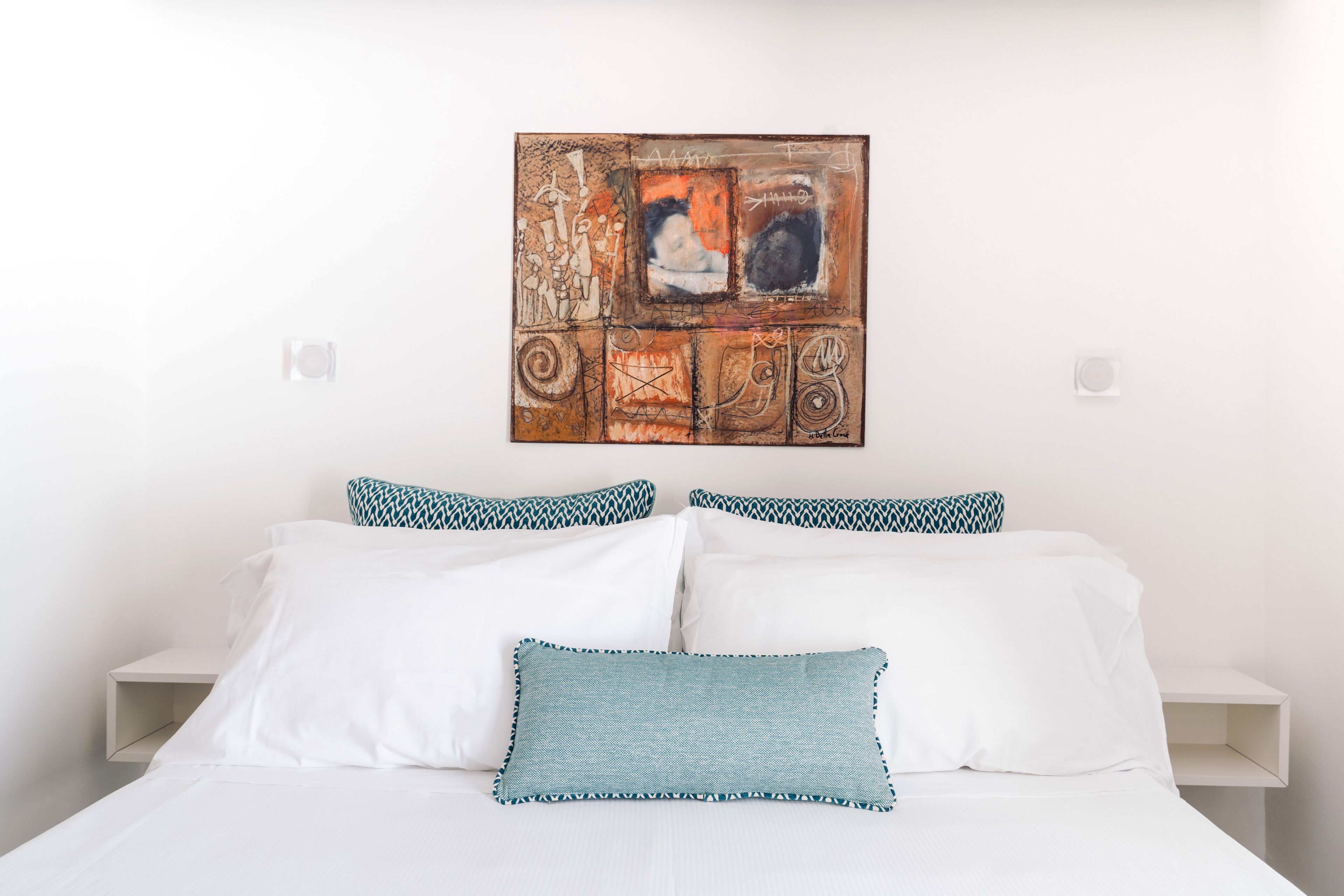 Una cama con sábanas blancas y almohadas estampadas, un cuadro abstracto colgado en la pared y una mesita de noche blanca.