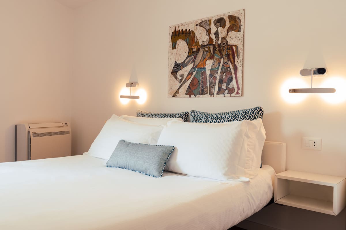 Un lit bien fait, avec du linge de lit blanc et un coussin bleu, un tableau abstrait accroché au mur, et deux tables de chevet avec des lampes murales.