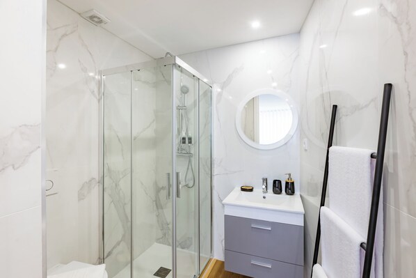 A modern bathroom with a glass-enclosed shower, a marble-tiled wall, a round mirror, and a vanity with a towel rack. - Apartment in the city center—modern, new, and cozy—with 2 bedrooms and 2 bathrooms (Porto)