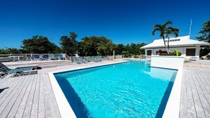 Una piscina rettangolare con un bordo in legno, un edificio bianco con una porta rossa e palme sullo sfondo.