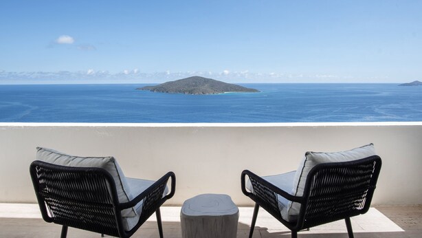 Un balcone con due poltrone in vimini e un tavolino rotondo, con vista su un mare tranquillo e un'isola all'orizzonte.