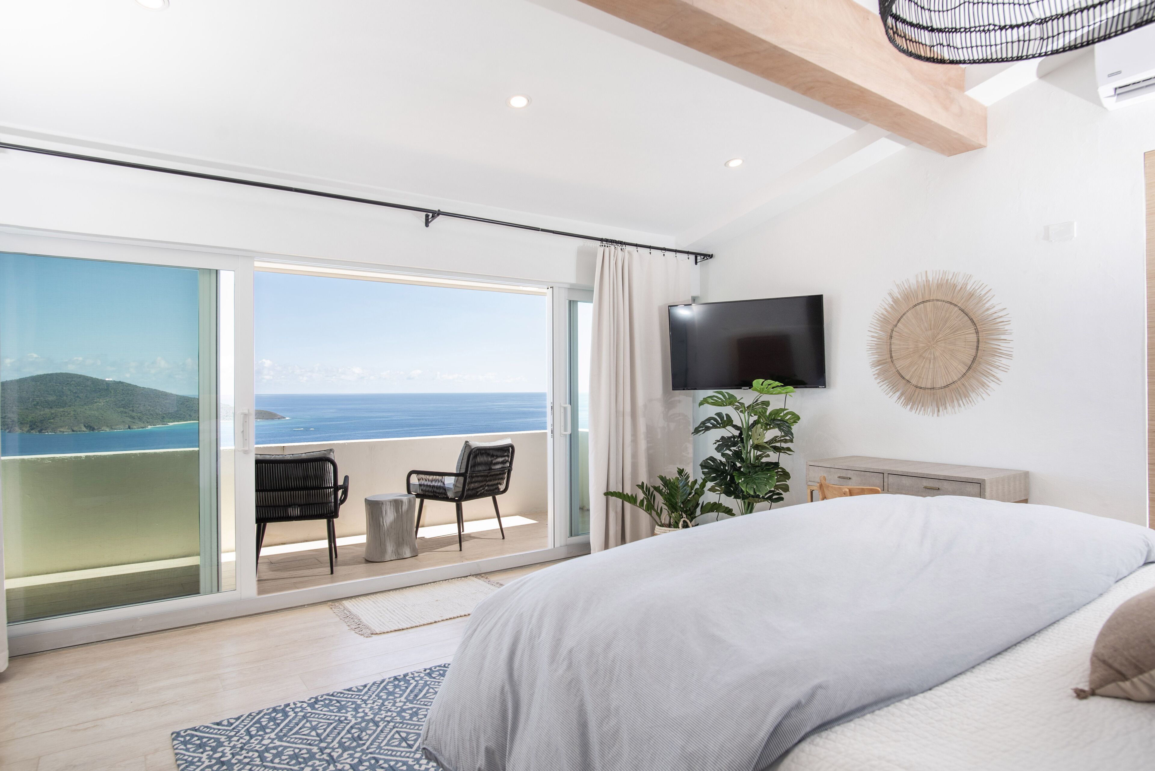 A bedroom with a large bed, a TV, a plant, and a view of the ocean.