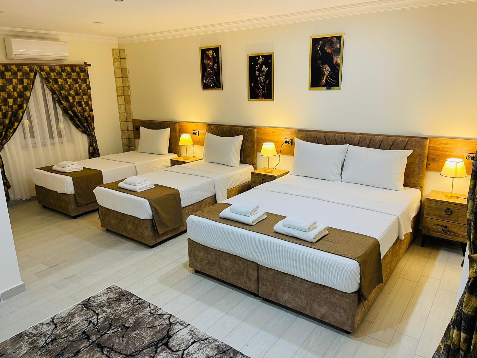 A hotel room with three single beds, a large bed, and a sofa bed. There are bedside tables with lamps, a window with curtains, and artwork on the wall.