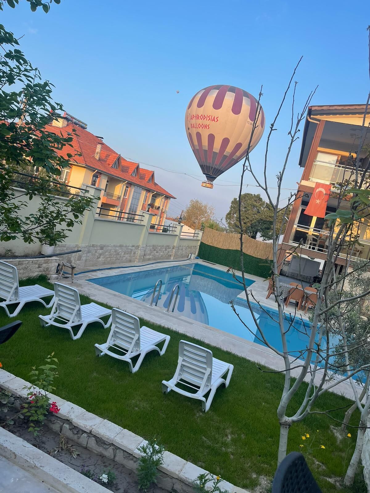 Una piscina con sillones de descanso, un globo aerostático al fondo y un edificio con tejado rojo.