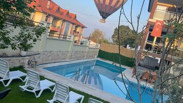 Una piscina con sillones de descanso, un globo aerostático al fondo y un edificio con tejado rojo.