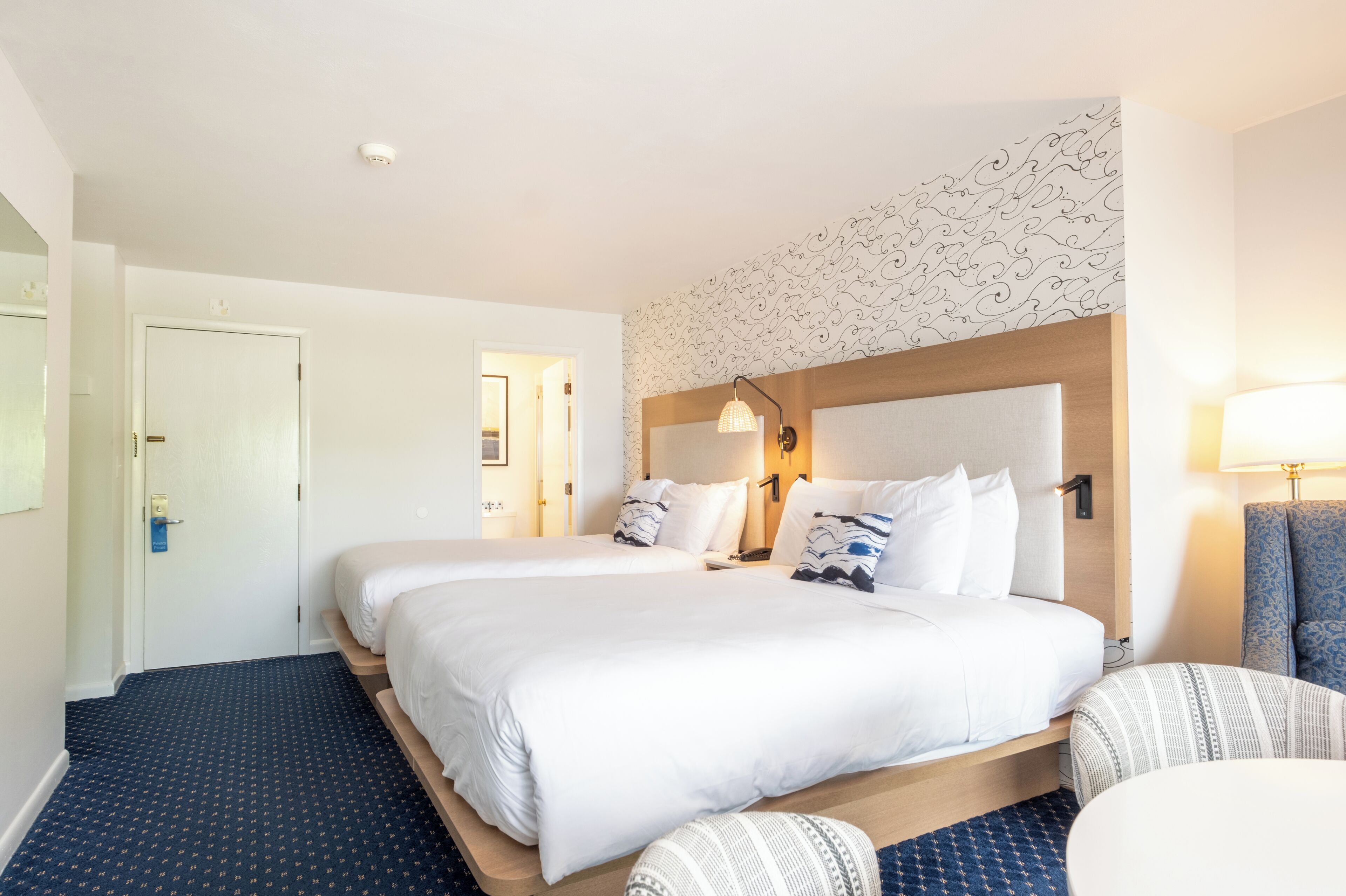 A hotel room with two beds, a wooden headboard, a patterned wall, a desk, and a chair.