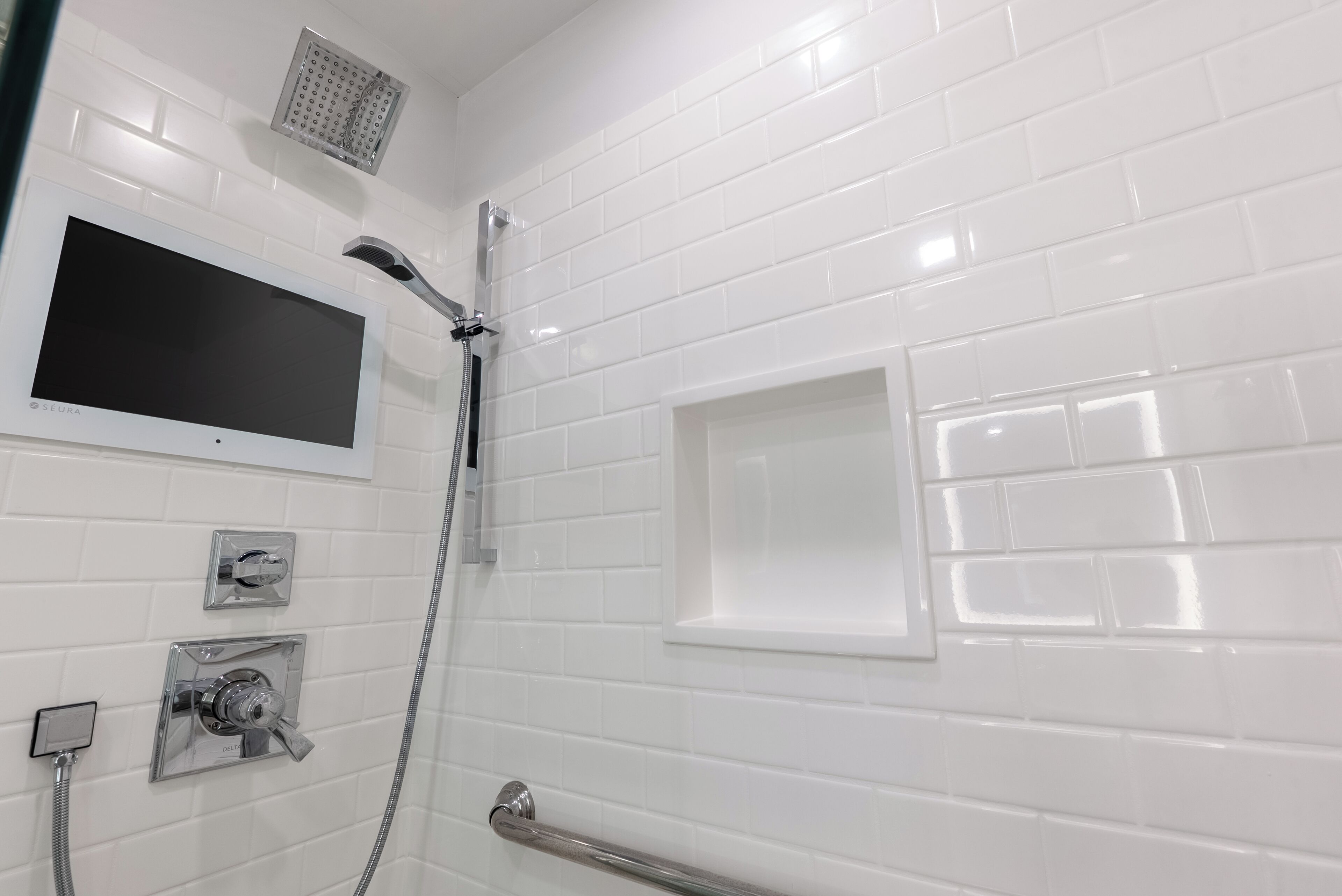 A bathroom with white tiled walls, a shower with a handheld showerhead, a handheld showerhead, and a glass-enclosed window.
