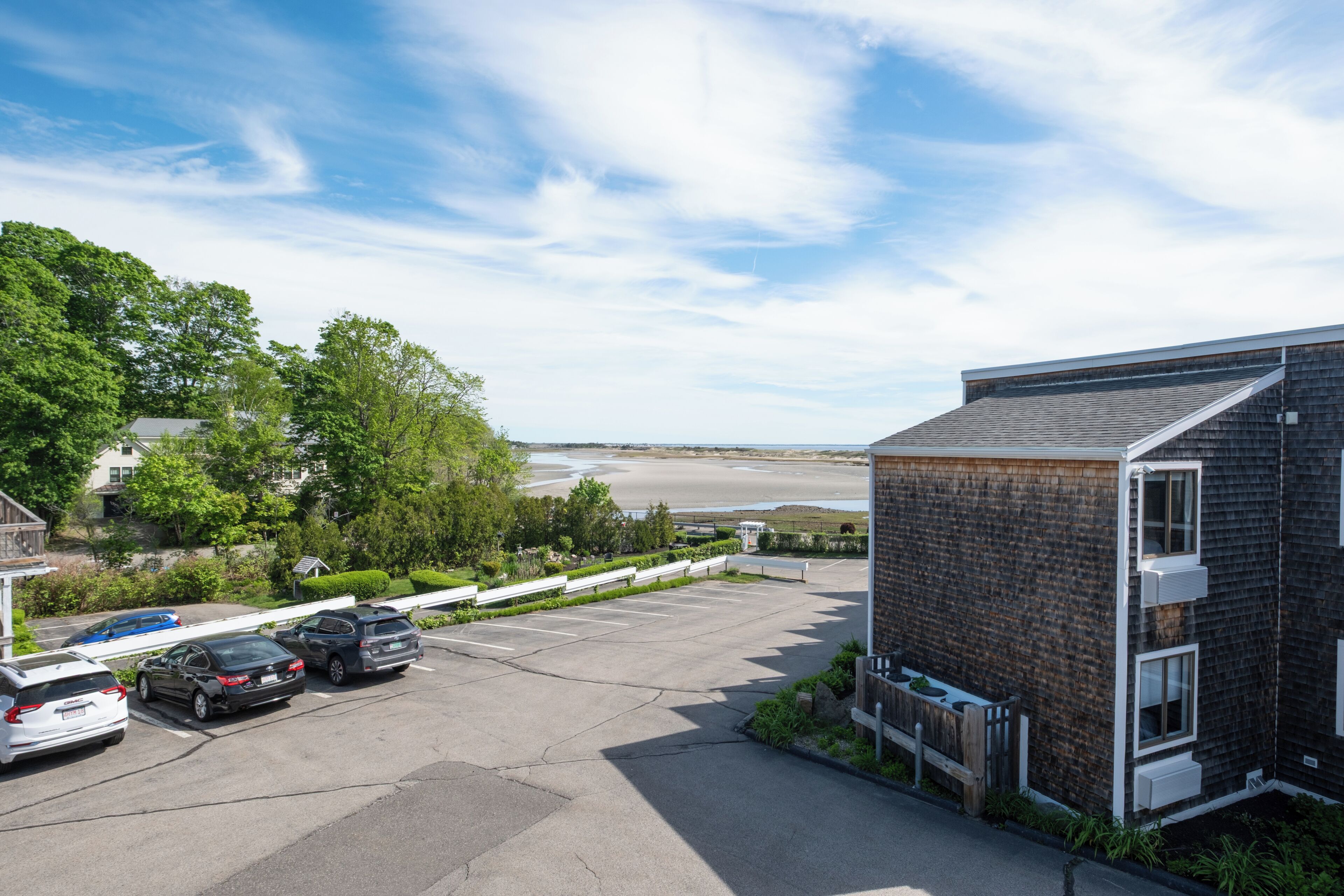 A parking lot with cars, a building with a sloped roof, and a view of a beach and water body.