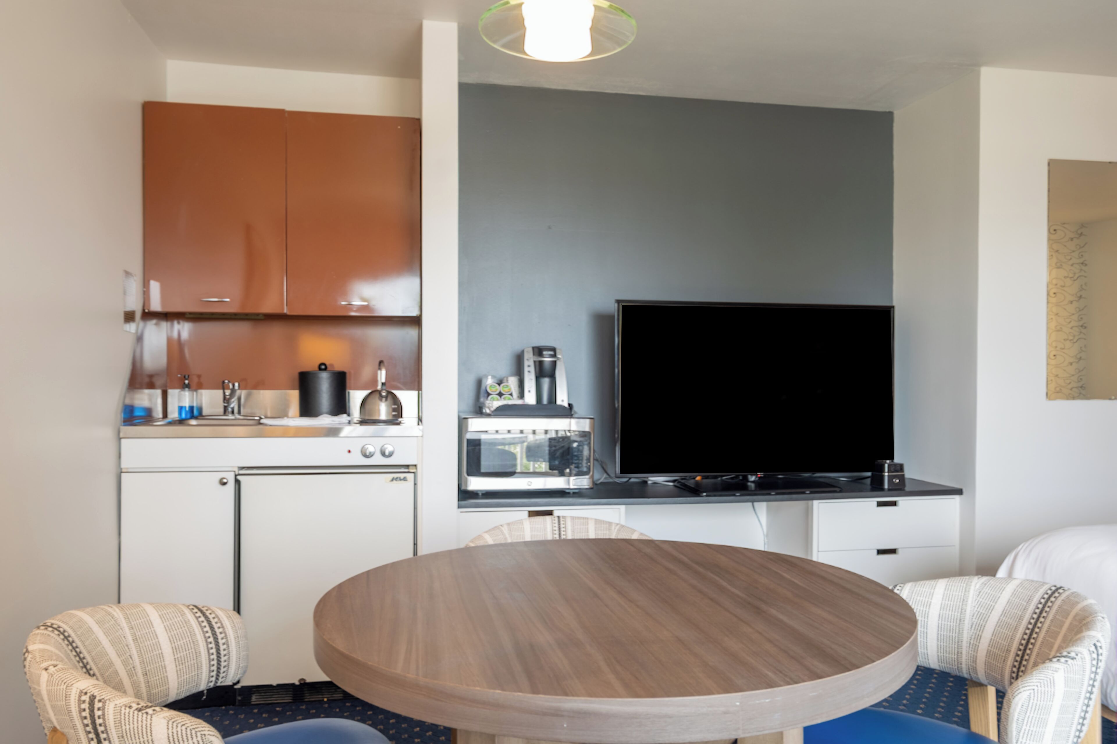 Cocina compacta con microondas, cafetera y televisor de pantalla plana. Hay una mesa redonda de madera con sillas y un refrigerador pequeño.
