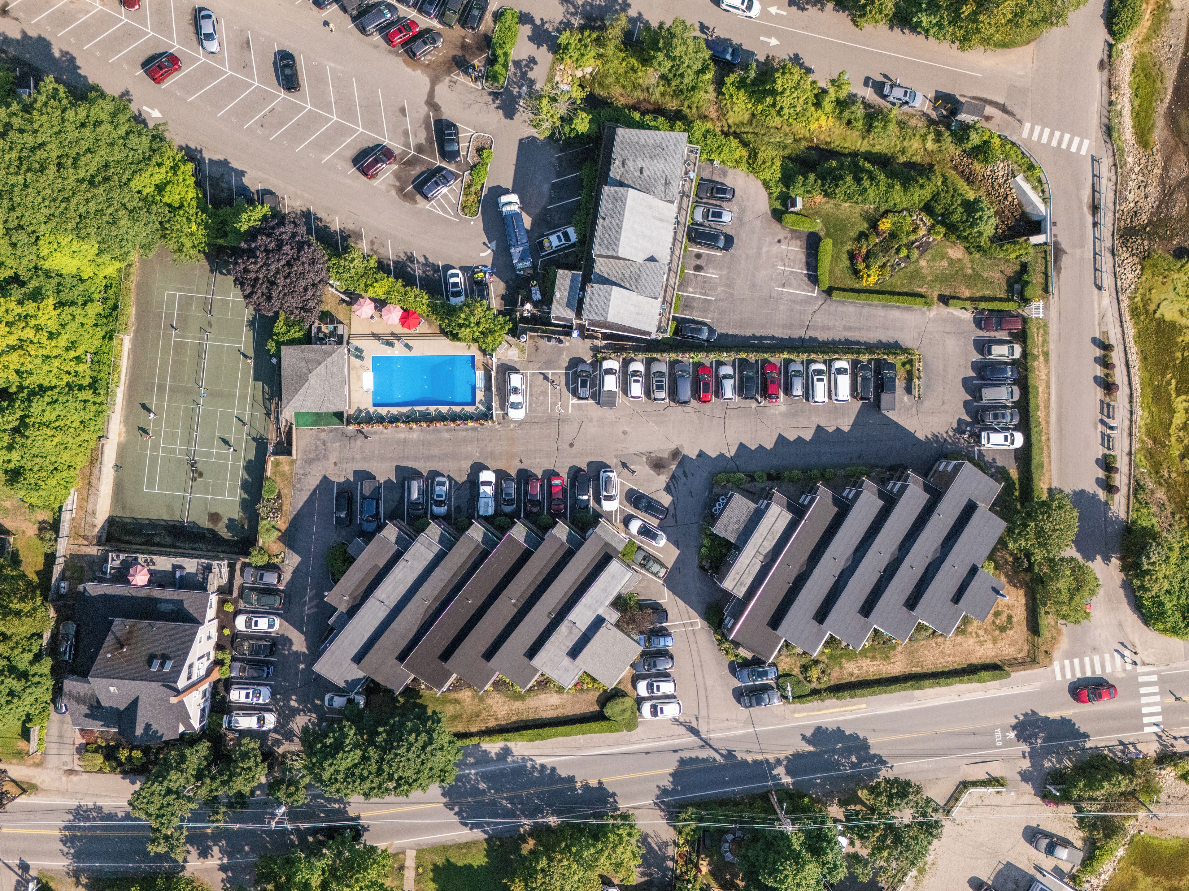 An aerial view of a parking lot with multiple rows of cars, a tennis court, and surrounding greenery.