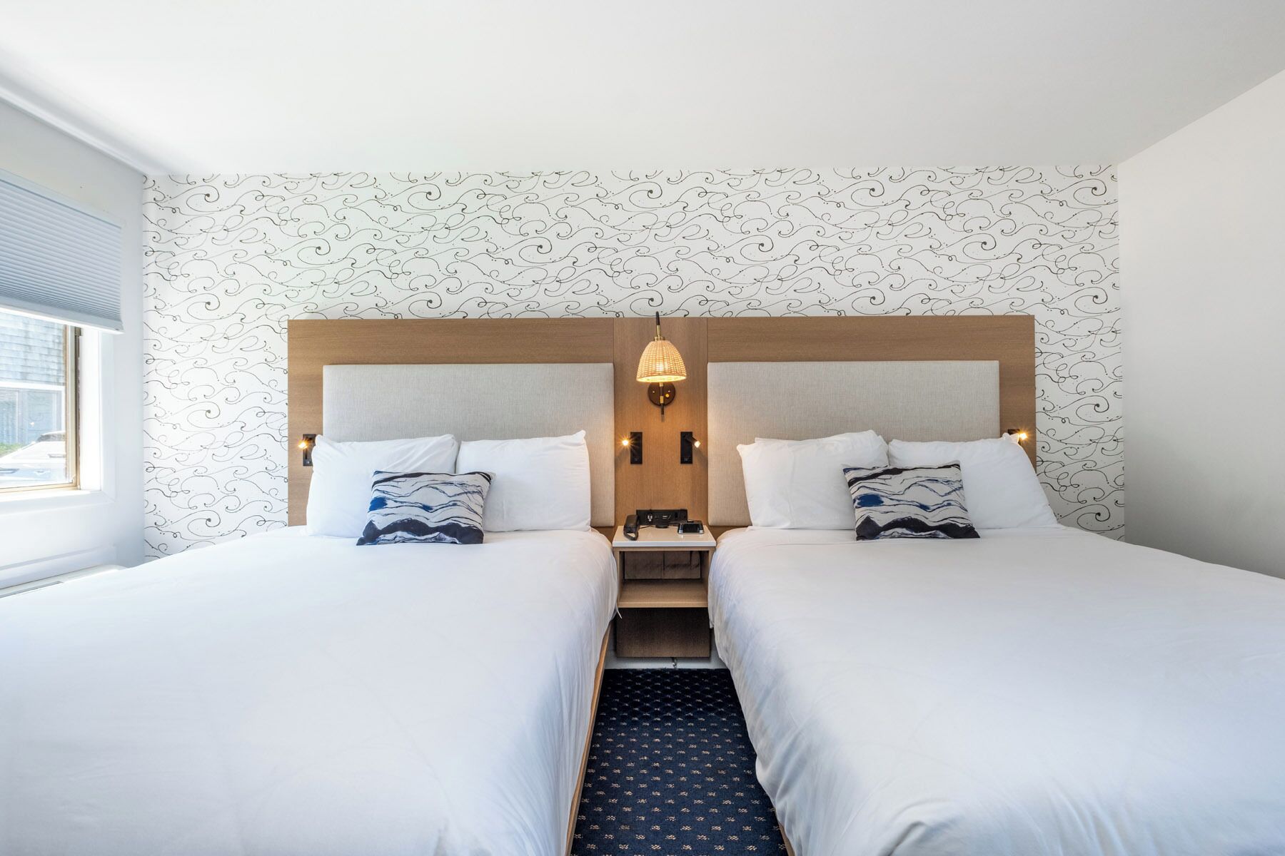 A double room with two beds, a wooden headboard, a nightstand with a lamp, and a patterned wallpaper.