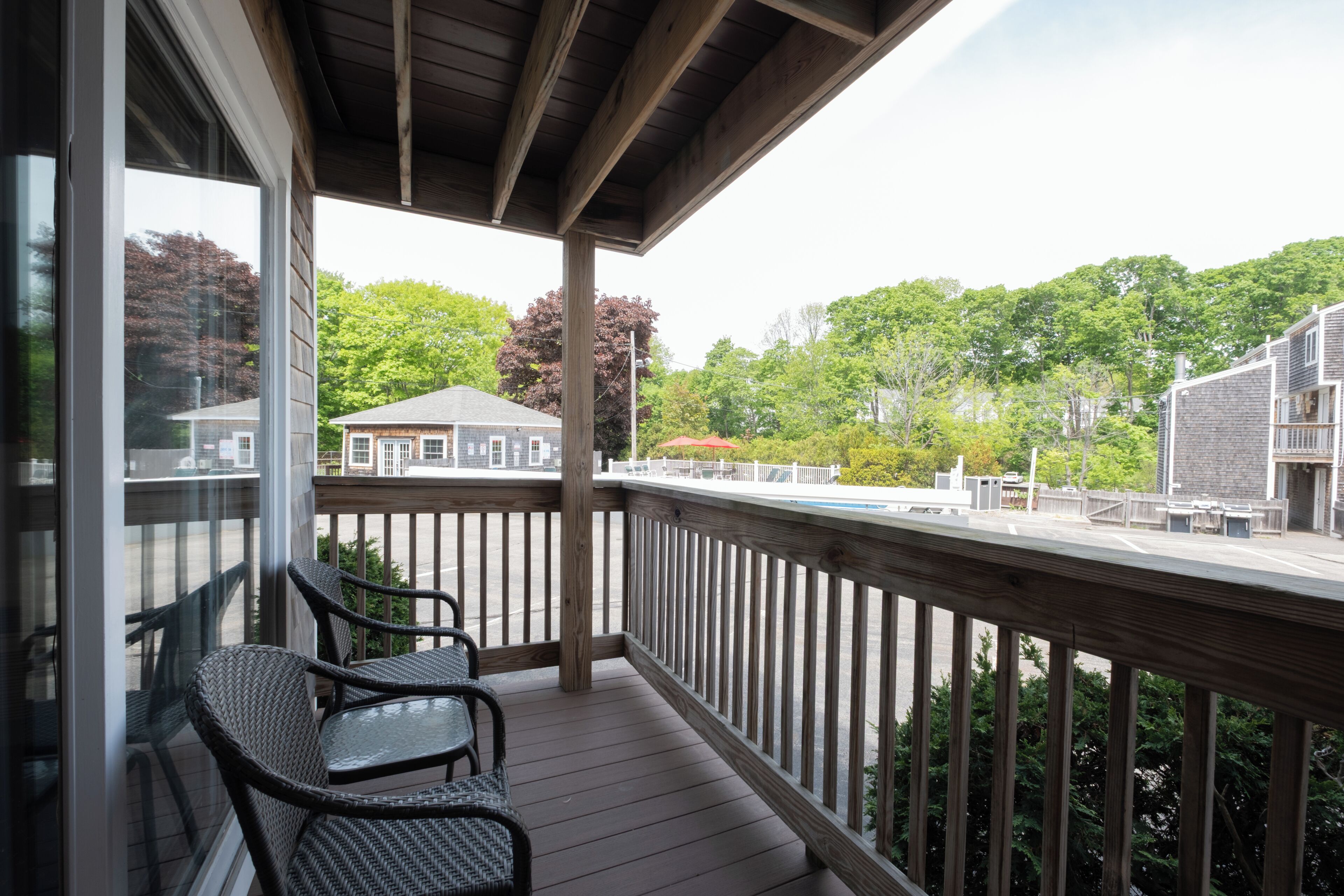 A covered balcony with wicker chairs, a wooden deck, and a view of a building and trees.