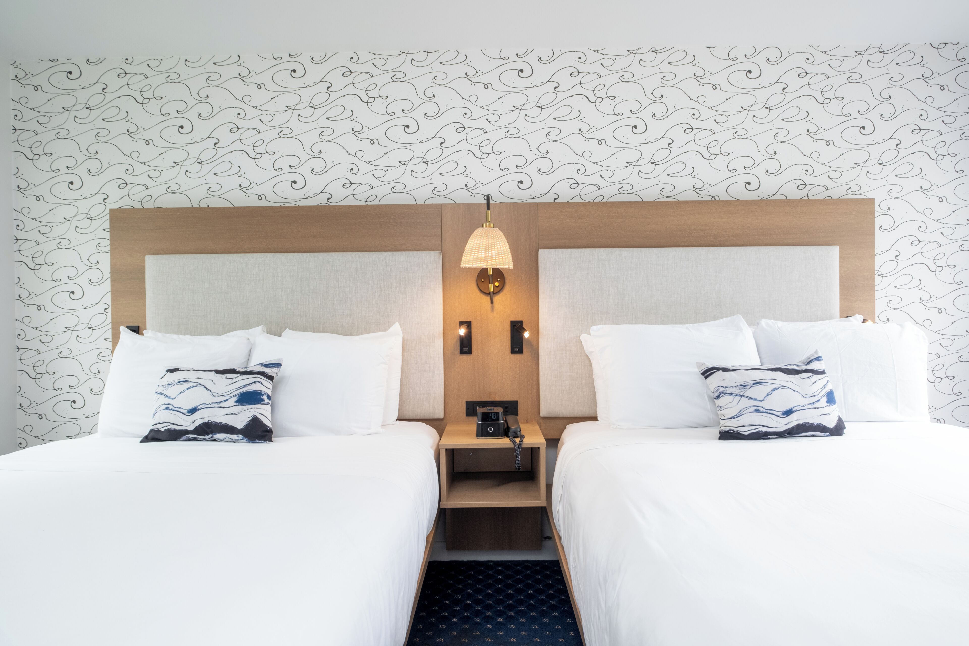 A hotel room with two beds, a nightstand, a lamp, and a patterned wallpaper.