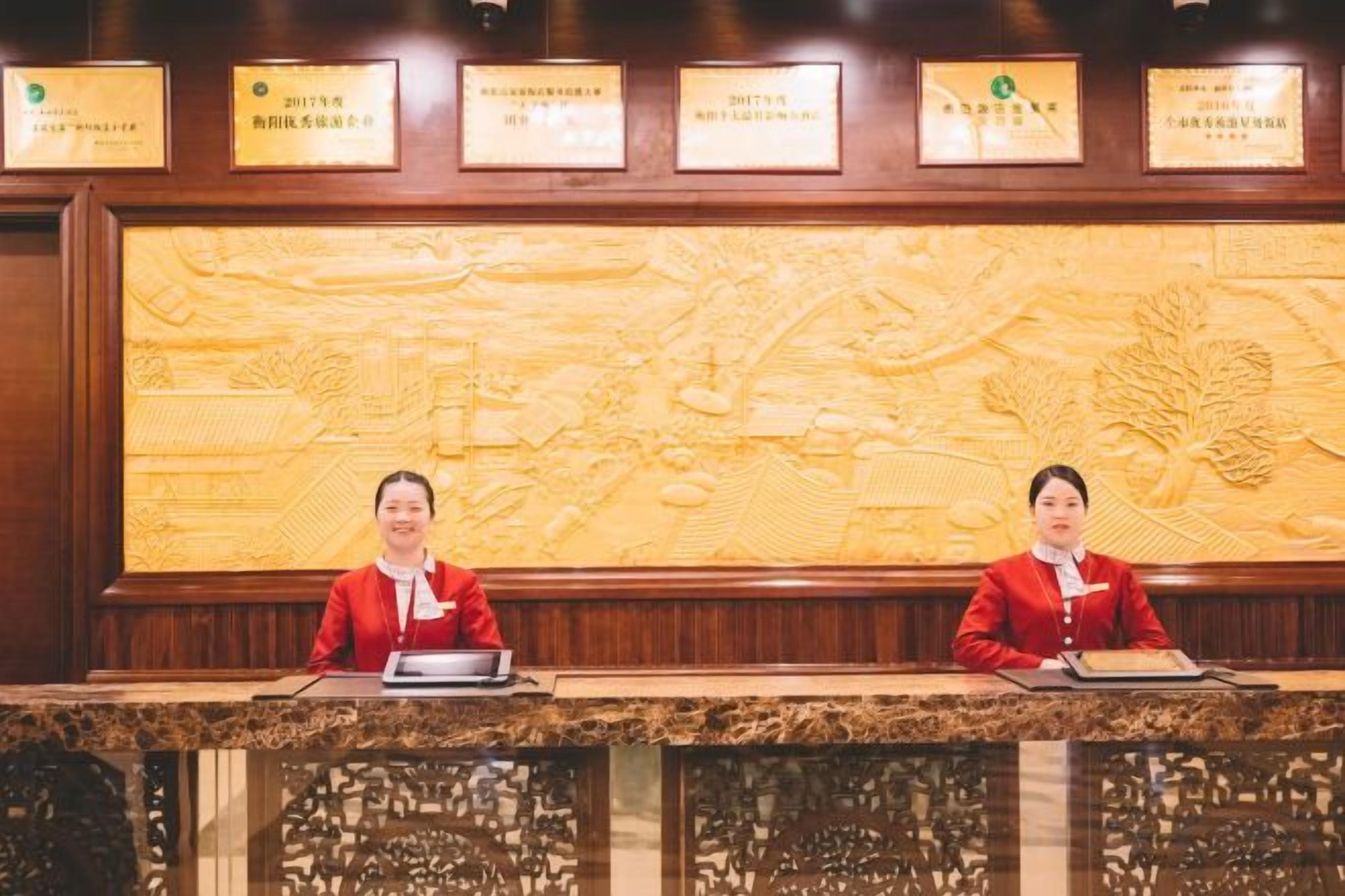 Un mostrador de recepción con dos recepcionistas, una gran pared tallada y certificados enmarcados arriba.
