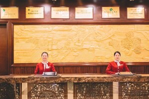 A reception desk with two receptionists, a large carved wall, and framed certificates above. - Shenlong New World Hotel (Leiyang)
