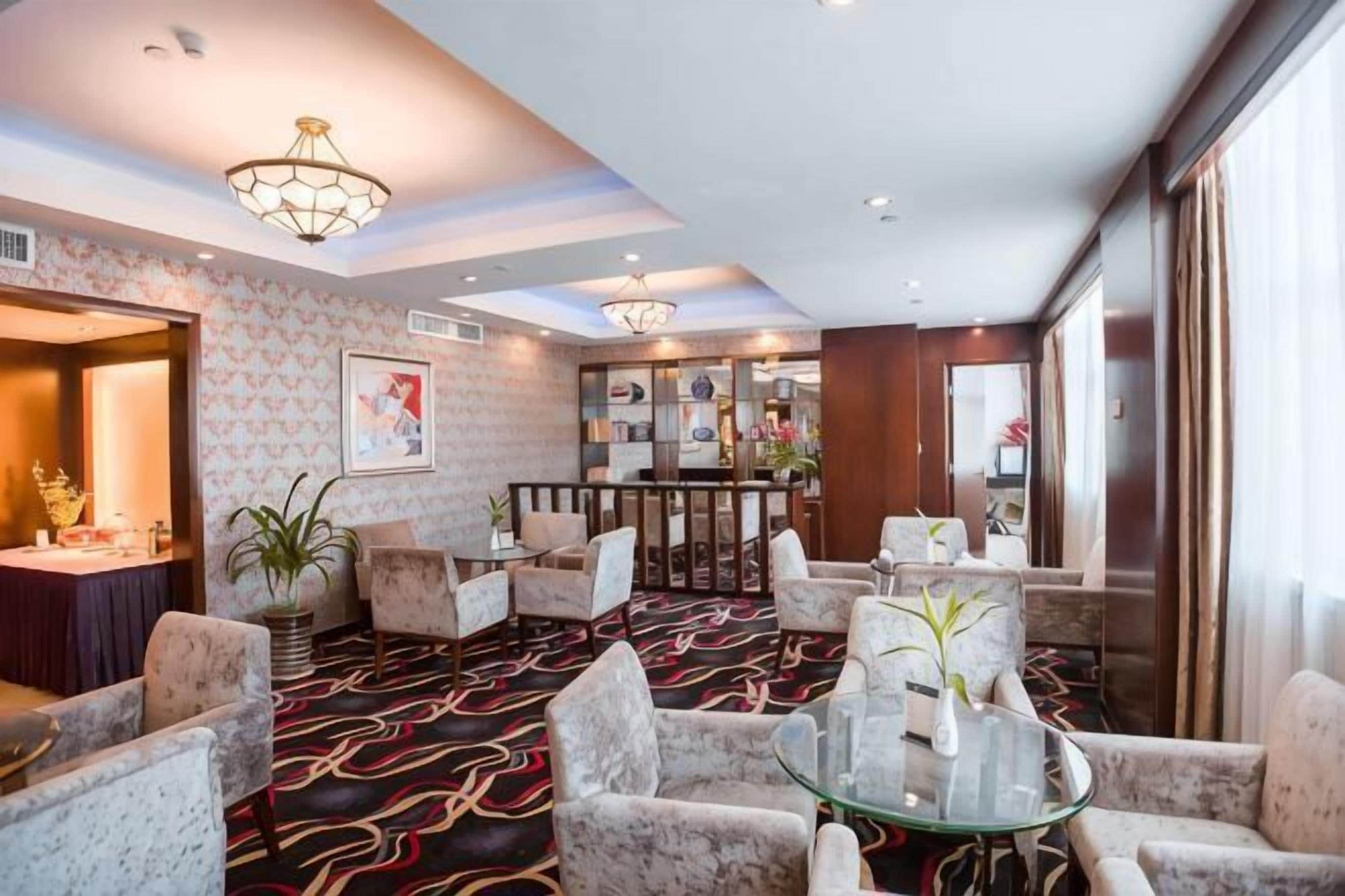 A hotel lobby with a patterned carpet, round tables, and chairs, a reception desk, and a bar area.