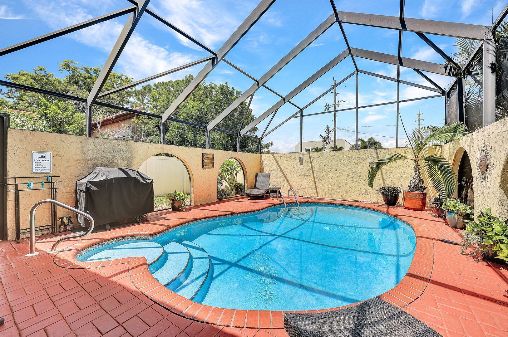 A swimming pool with a covered area, surrounded by brick flooring, and potted plants.
