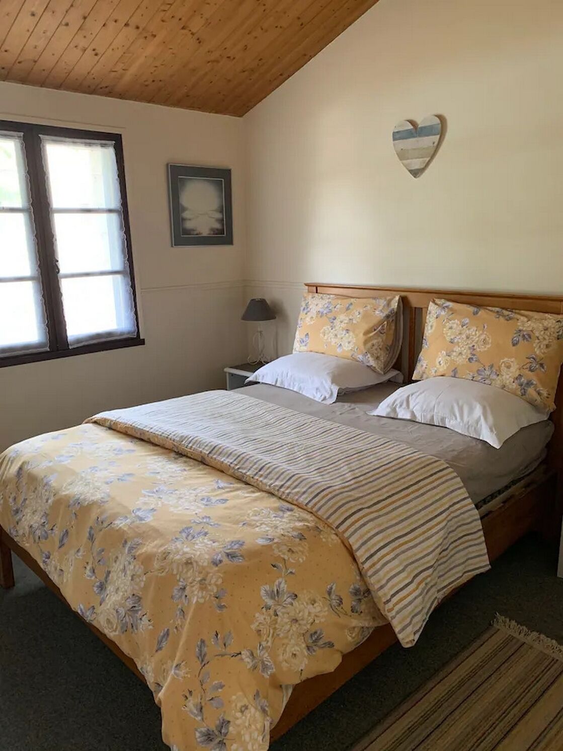 Une chambre avec un plafond en bois, un lit recouvert d’une literie à motifs floraux, une table de chevet avec une lampe et une décoration en forme de cœur accrochée au mur.