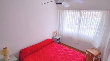 Un dormitorio con cama, mesita de noche, silla, ventilador de techo y ventana con cortinas.