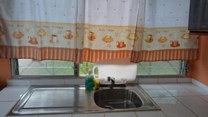Keittiössä on ruostumattomasta teräksestä valmistettu pesuallas, ikkunassa on kahvikuppikuviollinen verho ja työtaso on valkoinen.