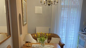 A living room with a brown sofa, a green armchair, a wooden coffee table with a vase of yellow flowers, a chandelier, and a kitchen area visible through a window with sheer curtains.
