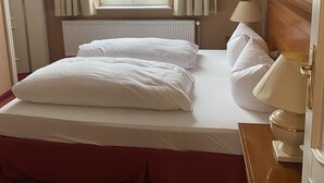 A hotel room with a large bed, a radiator, a window with curtains, a bedside table with a lamp, and a framed picture on the wall.