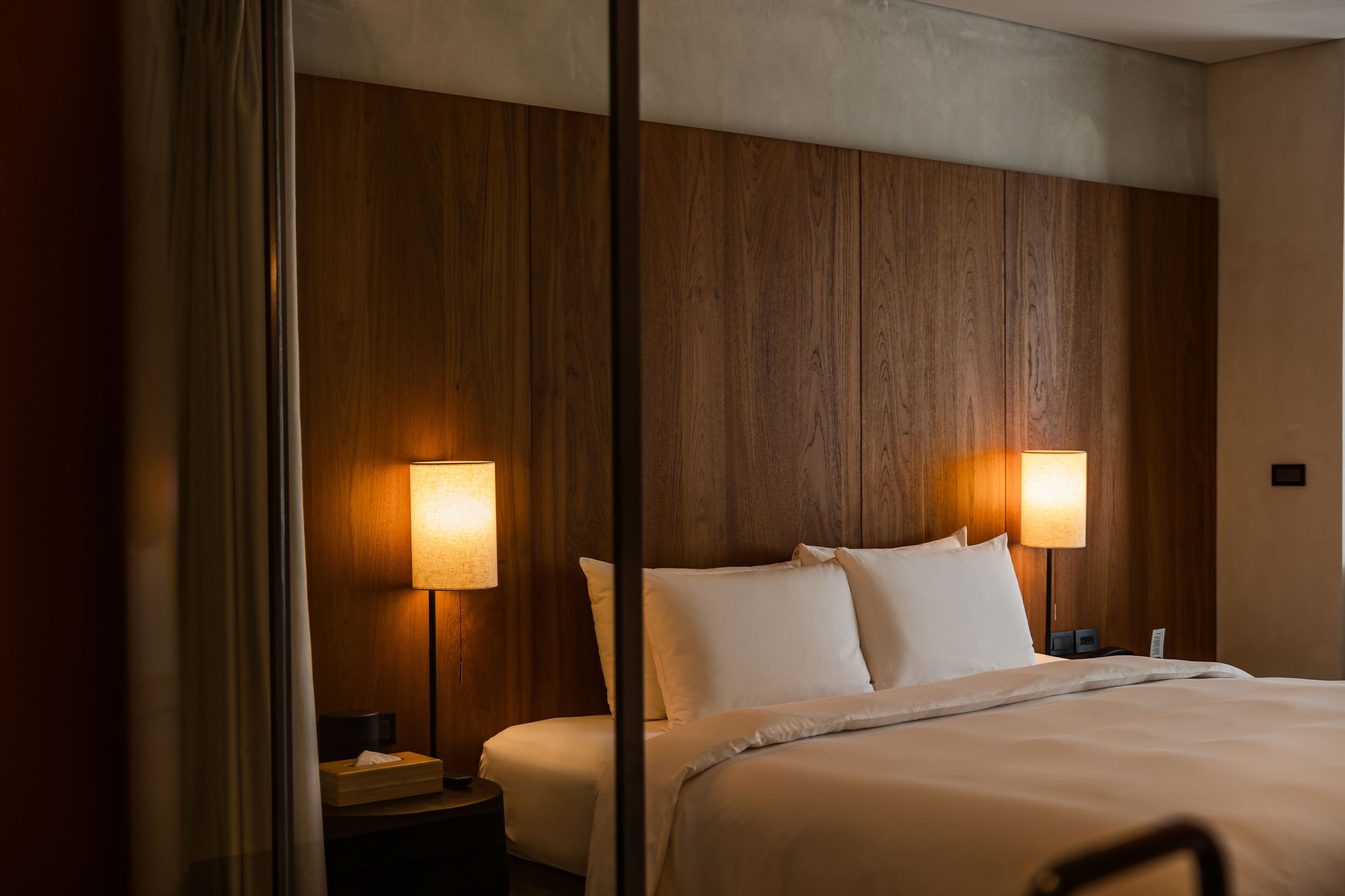 Une chambre d’hôtel avec une tête de lit en bois, deux lampes de chevet, un lit avec du linge de lit blanc et une table de chevet avec un téléphone et une boîte de mouchoirs.